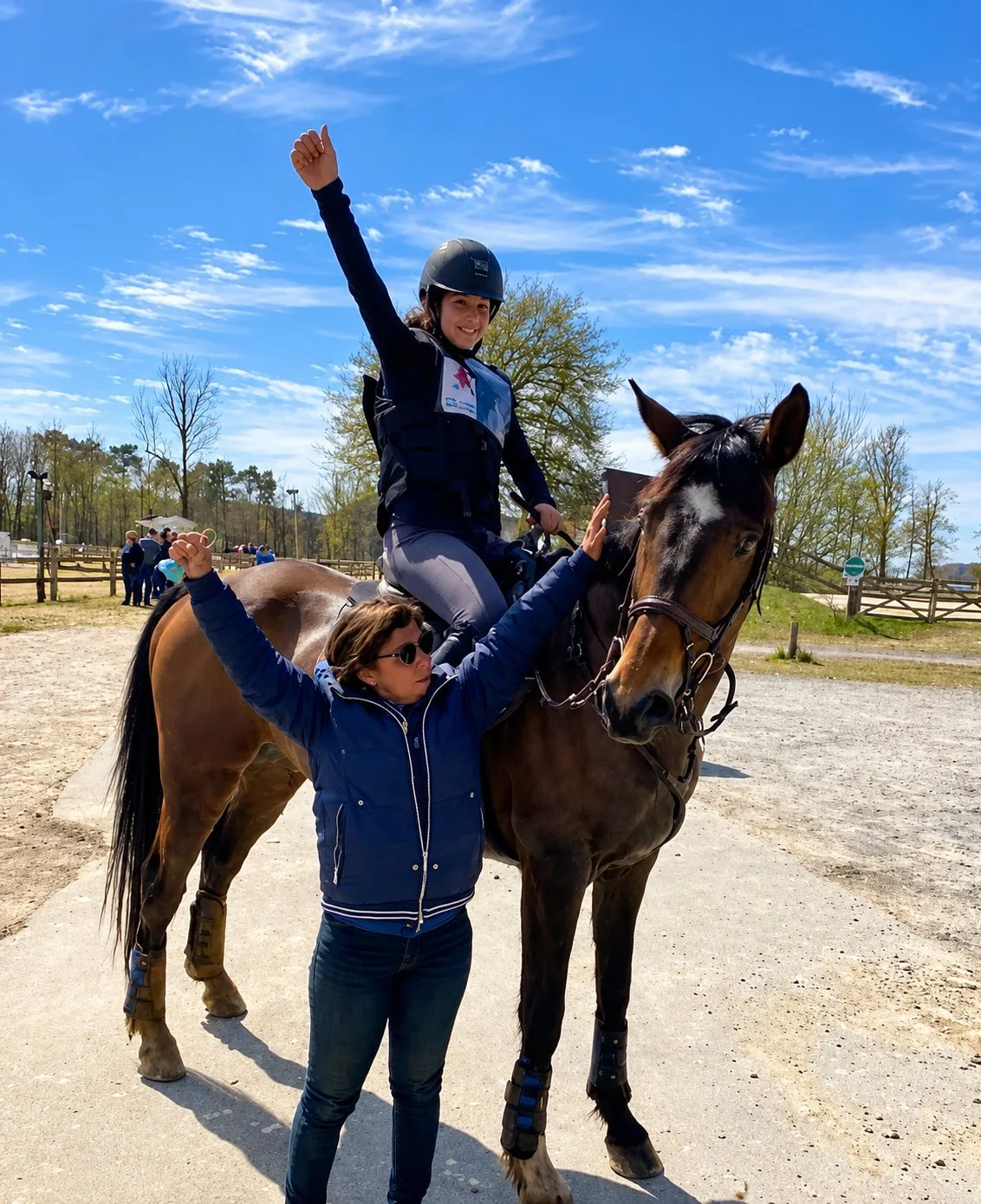 Une cavalière en casque lève le bras en selle sur un cheval alezan, tandis qu'une femme en doudoune bleue se tient devant lui dans une carrière sous un ciel bleu.