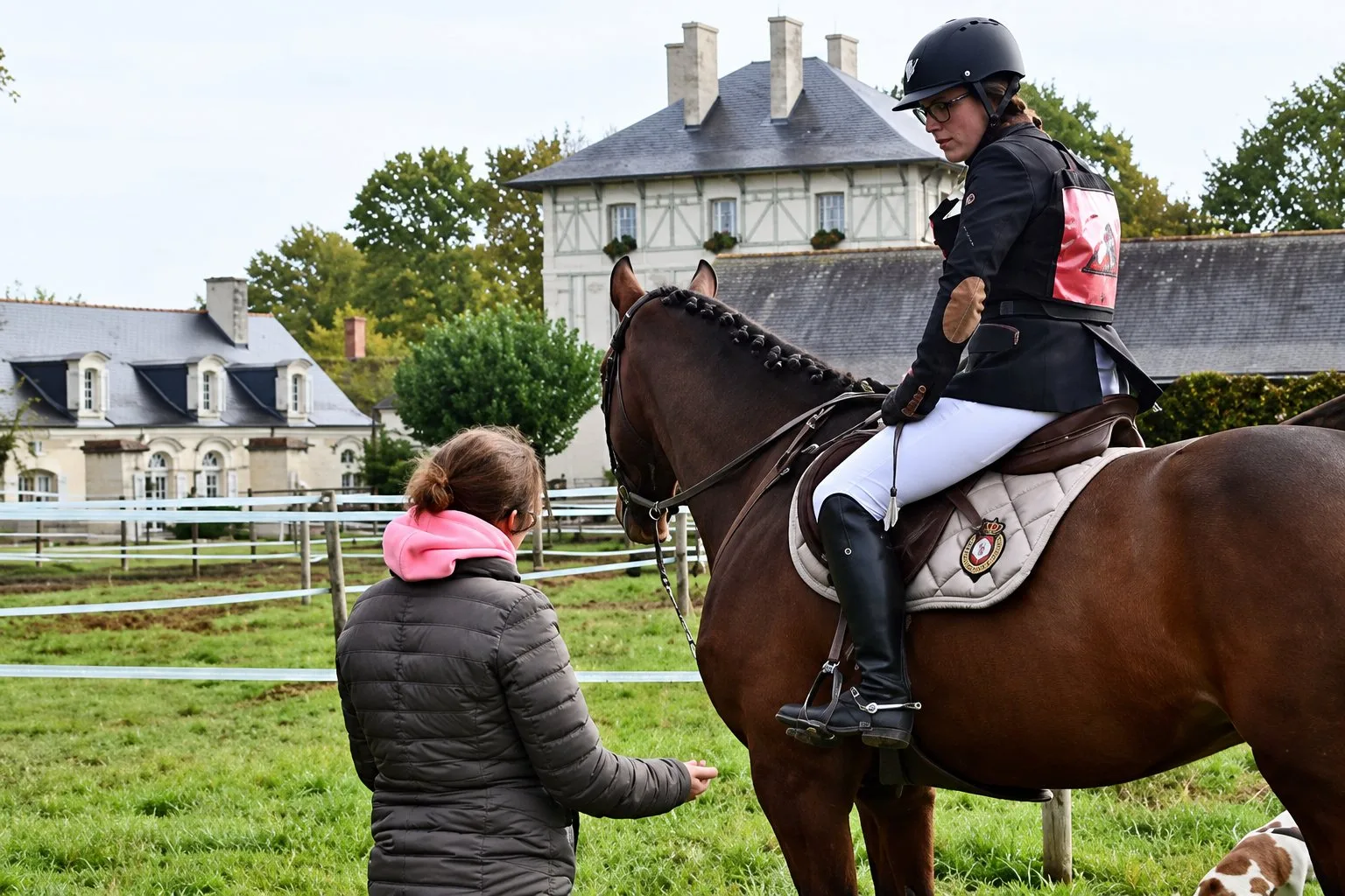 Une cavalière en tenue noire et pantalon blanc, casque sur la tête, monte un cheval alezan pendant qu'une femme en doudoune lui parle, devant des bâtiments en pierre.