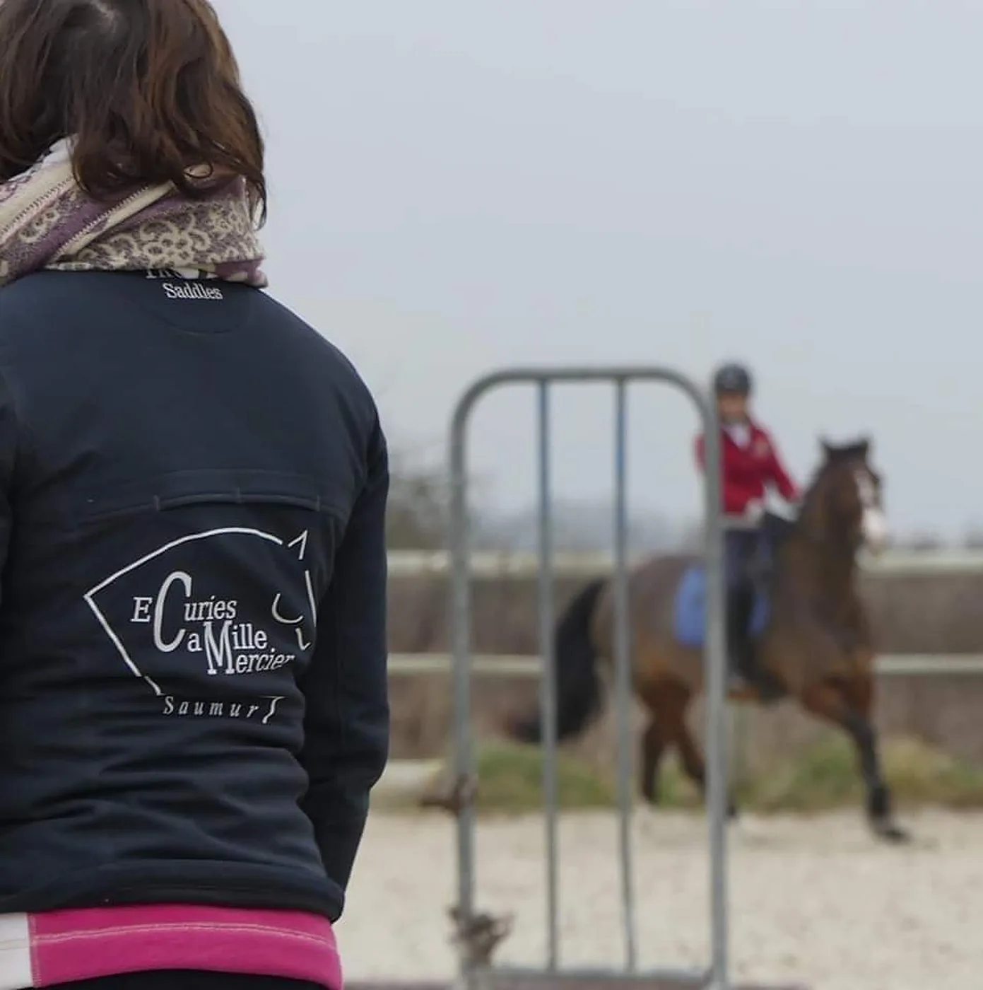 Une personne vue de dos portant une veste marquée du nom d'un club équestre de Saumur regarde une cavalière en veste rouge monter un cheval alezan dans un manège en sable.