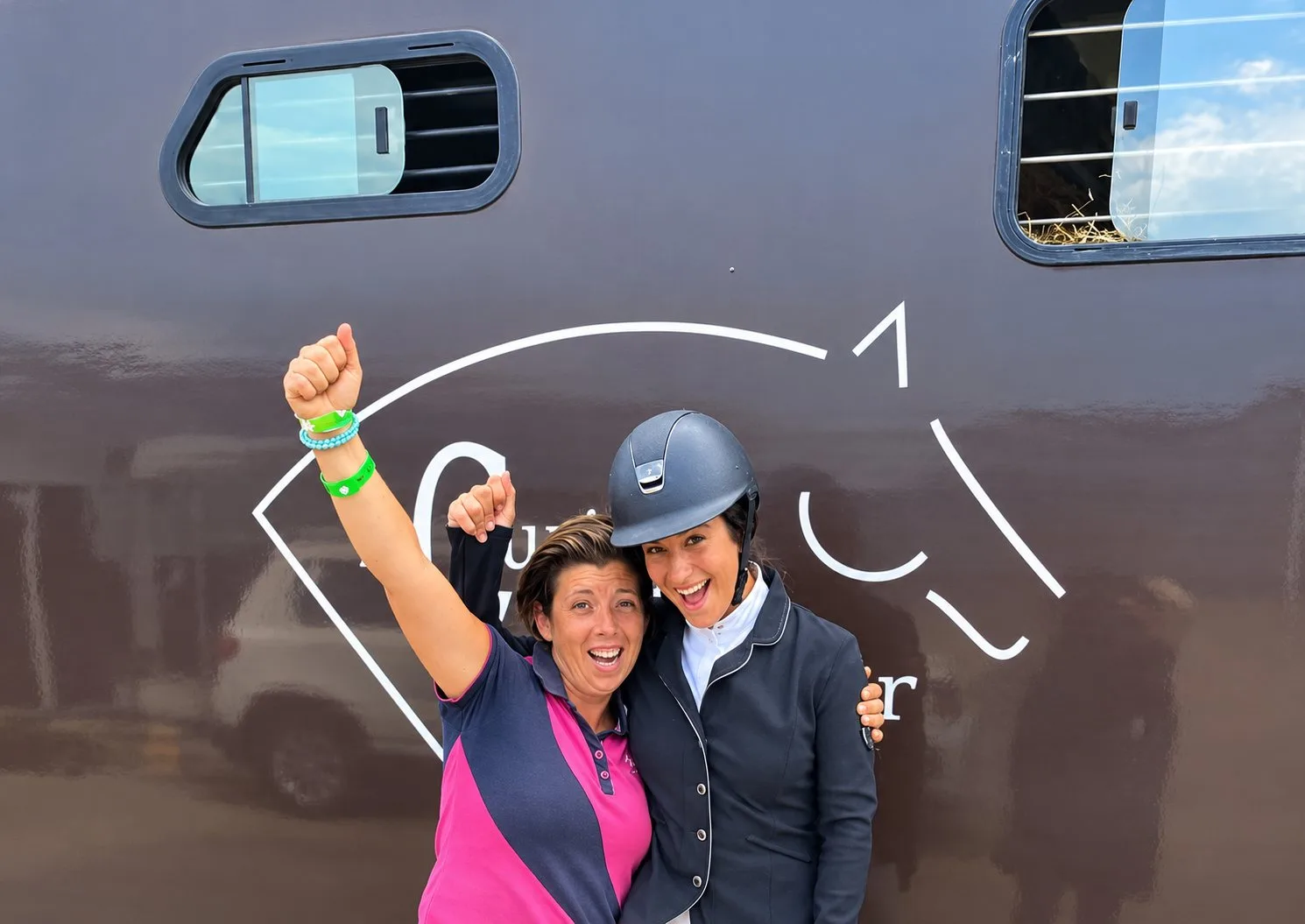 Deux femmes souriantes posent en levant le poing devant un van marron orné d'une tête de cheval stylisée, l'une portant un casque d'équitation.