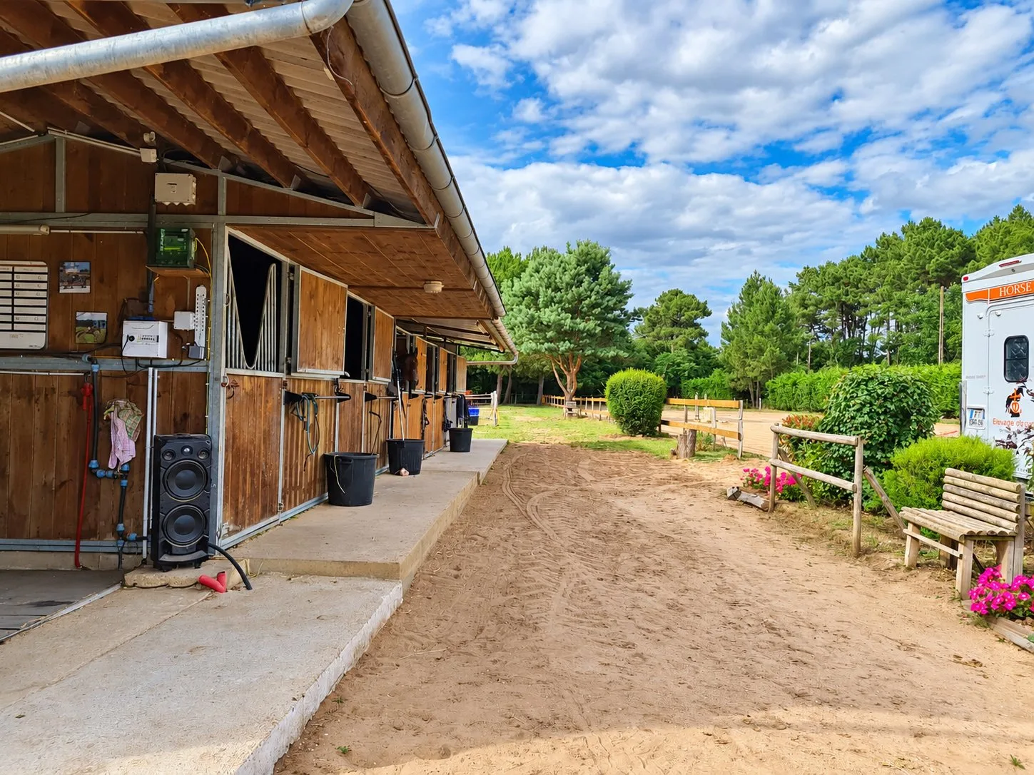 Une rangée de boxes en bois sous un auvent borde une allée de sable, avec un banc, des fleurs roses et un van à droite sous un ciel nuageux.