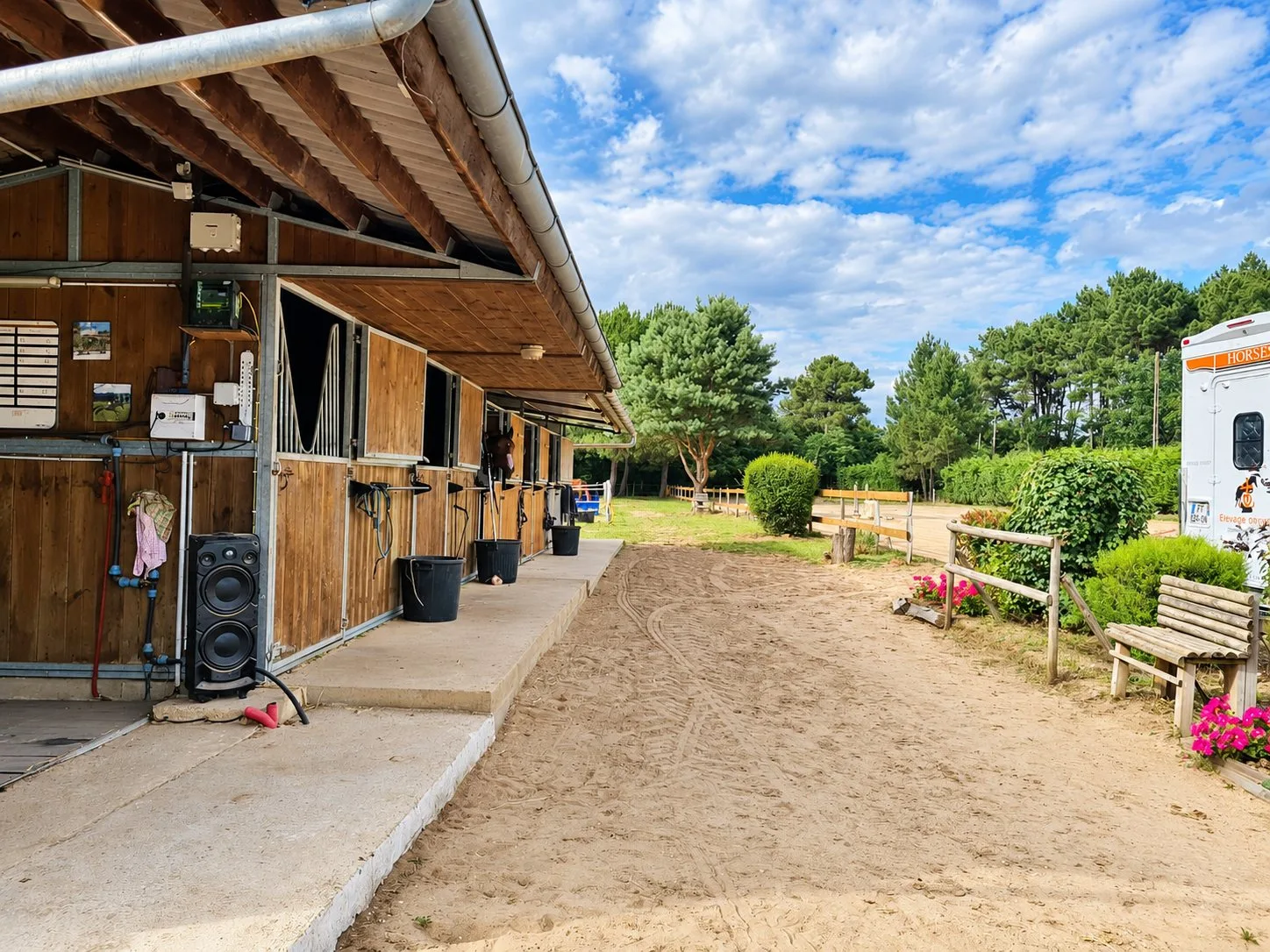 Une rangée de boxes en bois sous un auvent borde une allée de sable, avec des massifs de fleurs roses, un banc en bois et des pins en arrière-plan.