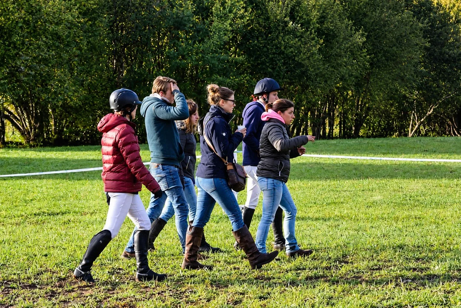 Un groupe de cavaliers en bottes et vestes, certains portant un casque, marche sur l'herbe le long d'une bande blanche devant une rangée d'arbres.