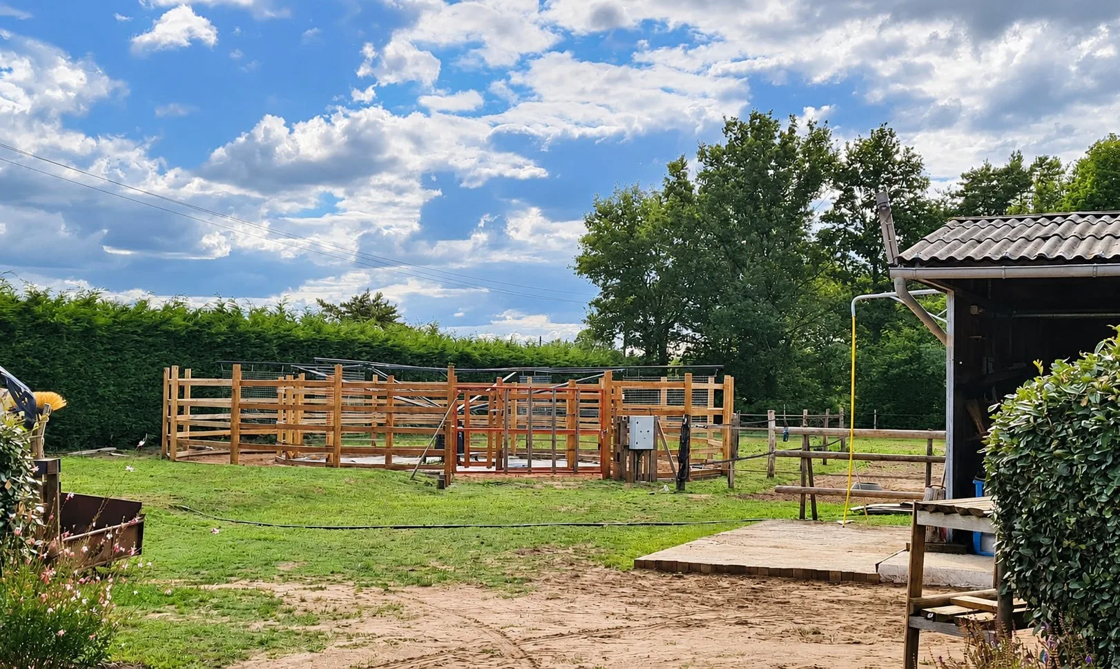Une grande structure en bois à claire-voie, pour chevaux, est installée sur l'herbe devant une haie, à côté d'un bâtiment sombre et d'un chemin de sable.